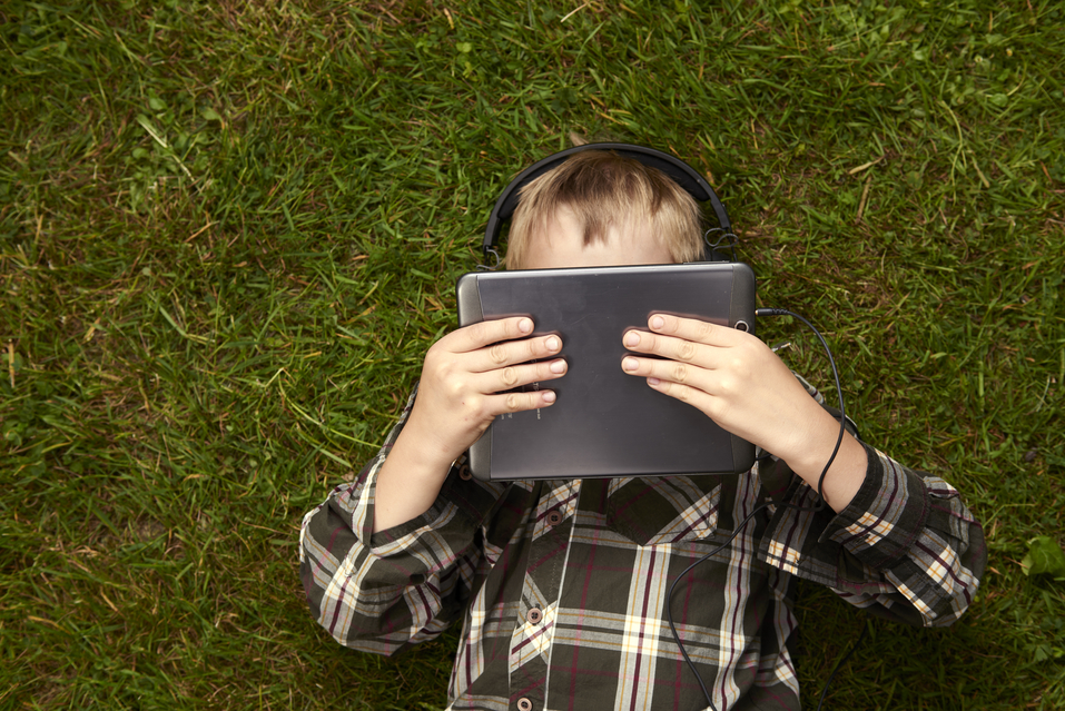Portrait of Child blond young boy playing with a digital tablet pc computer outdoors  lying on grass, listening with headphones