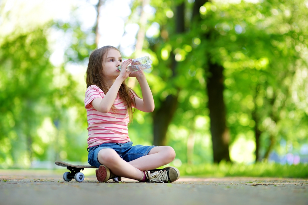 Pretty little girl drinking water while sitting on a skateboard outdoors on beautiful summer day