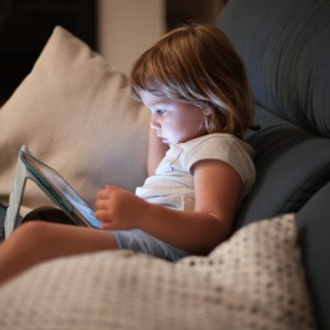 blonde three years old baby shirt and shorts, sitting comfortably in sofa inside home at night reading and watching digital tablet, face illuminated by the light of the screen